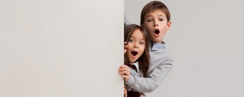 Banner with a surprised children peeking at the edge with copyspace. The portrait of cute little kids boy and girls looking at camera against white studio wall. Kids fashion and happy emotions concept