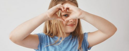 Close up of beautiful blonde girl making heart with hands, looking through it in camera, posing for photo with smile and happy expression