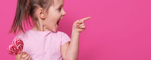 Close up portrait of little attractive child opening mouth widely, looking other side with excitement, holding heart bright lollipop. Playful merry little fair haired girl spends sparetime happily.